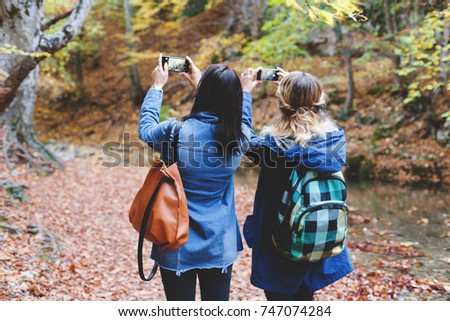 Two young girl friends walking in an autumn forest and taking pictures of the beautiful nature on the smartphone. Walk in the woods. Girlsfriends hiking at fall.