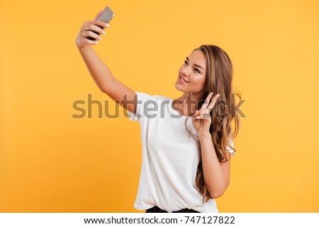 Photo of pretty cute beautiful young woman make selfie by mobile phone isolated over yellow wall background. Looking aside showing peace gesture.