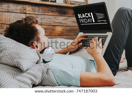 Black Friday banner in a laptop computer while man uses it to buy by internet lying down at home.