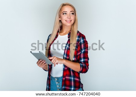 Working with joy. Beautiful young woman with blond hair holding her digital tablet and
looking away with smile while standing against grey background