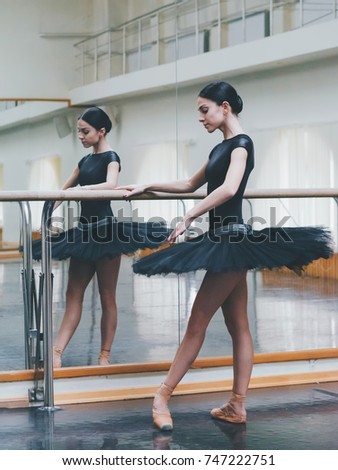 Ballerina in black tutu and pointe stretches on barre in ballet gym. Woman standing near bar and mirror, preparing for perfomance.