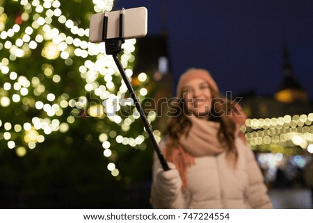 holidays and people concept - beautiful happy young woman taking picture by smartphone on selfie stick at christmas tree and market in winter evening