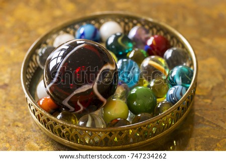 Large dark red glass shooter marble placed on top of smaller player marbles in small dish on golden background
