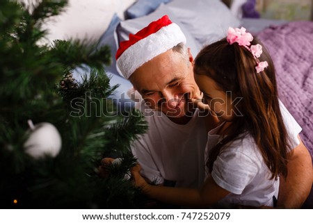 Picture of an elderly man wearing a Santa hat while playing with his grandchildren near a Christmas tree