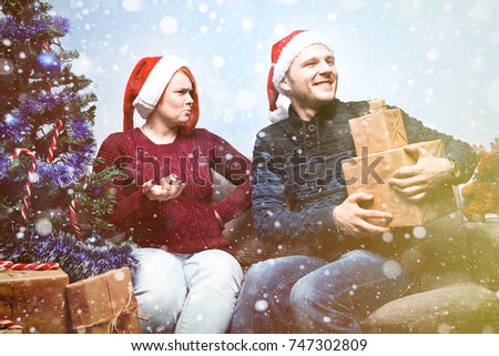 Spirit of Christmas and new year. Concept of a holiday and days off. A loving couple in Santa Claus hats. The girl is indignant with a small gift, the guy enjoys great gifts. with snowfall