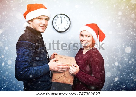 Spirit of Christmas and new year. Concept of a holiday and days off. A loving couple in Santa Claus hats. A girl and her boyfriend hold a gift at midnight. with snowfall