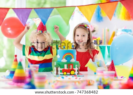 Kids birthday party. Child blowing out candles on colorful cake. Decorated home with rainbow flag banners, balloons, confetti. Farm and transport theme. Little boy celebrating birthday. Party food.