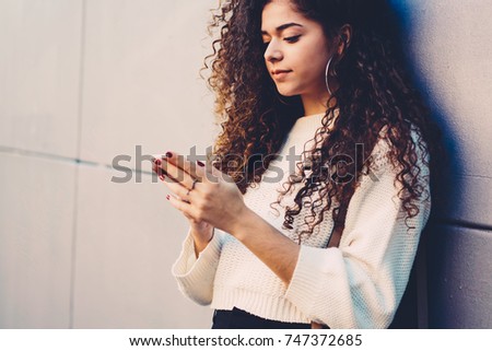Cropped image of young female person with beautiful curly hair watching interesting video in website on telephone outdoors.Pondering hipster girl checking account balance on telephone outside