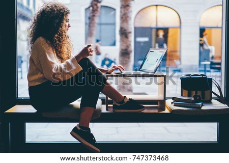 Young pondering female person with beautiful curly hair checking account balance on webpage.Talented student preparing for upcoming examinations during sitting next to copy space for your advertising Royalty-Free Stock Photo #747373468