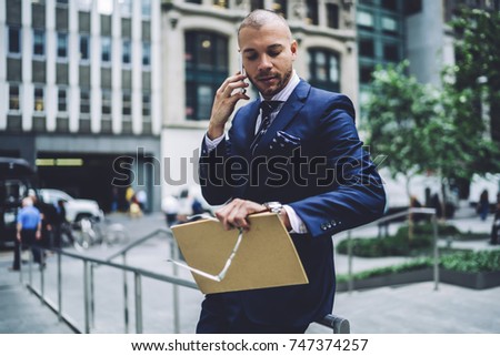 Serious executive manager talking on mobile planning meeting checking time standing on urban setting background, confident male lawyer looking at watch during mobile conversation with colleague 
