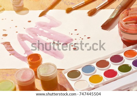 Background image showing interest in watercolor painting and art. A painted sheet of paper, surrounded by brushes, jars of watercolor paint and gouache, which lie on an old and stained wooden surface