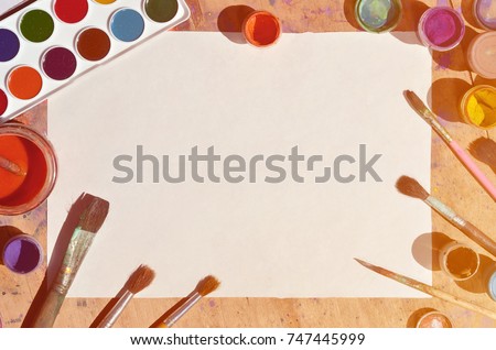 Background image showing interest in watercolor painting and art. A blank sheet of paper, surrounded by brushes, cans with watercolor paint and gouache, which lie on an old and stained wooden table