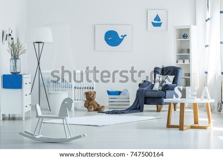 White rocking horse and table in spacious child's room with vase on cupboard, lamp and plush toy near armchair