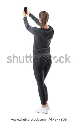 Rear view of sporty athletic woman taking selfie while holding cellphone in both hands. Full body length portrait isolated on white background. 