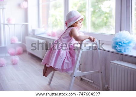 Sad baby girl in a pink dress and hat looks in the big window