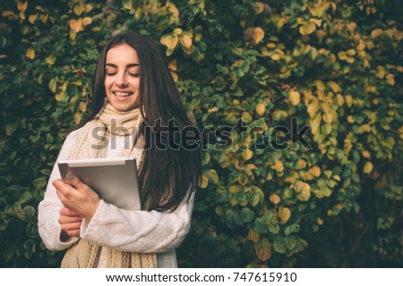 Beautiful young brunette sitting on a fallen autumn leaves in a park, reading an e-book on a tablet computer