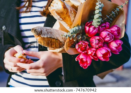 Young beautiful girl brunette in red beret and black coat goes along the street of the European city with bouquet of flowers and French baguettes. happy smile and charming look, French style of Paris Royalty-Free Stock Photo #747670942