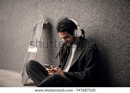 Stylish young dreadlock hipster with headphones sitting leaning against the grey wall and skate near him looking on a mobile.