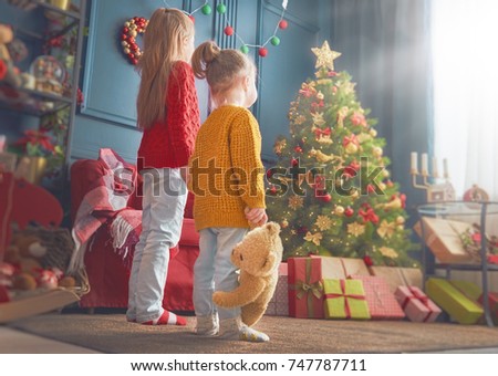 Merry Christmas and Happy Holidays! Cheerful cute little children girls. Kids sisters near tree indoors.
