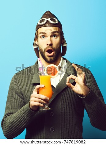 handsome bearded aviator man with long beard on surprised face holding glass of alcoholic cocktail in gray knitted sweater with hat and glasses on blue studio background