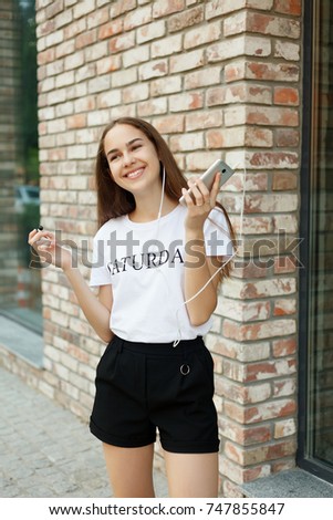 technologies, emotions, people, music, beauty, fashion and lifestyle concept - Young woman with headphones dangling on her mobile phone as she walks in an urban street, view from a height