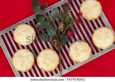 Fresh golden frosted mince pies on a cooling rack with a christmas acorn and berry arrangement against a red cloth background