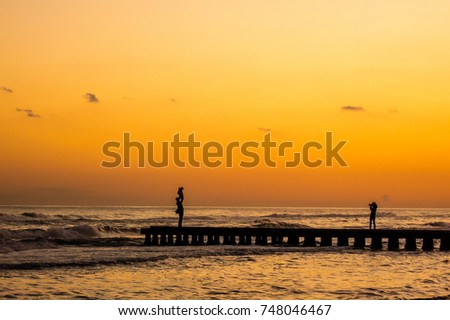 a woman photographs a dad that raises her son in the air, the scene is a silhouette takes place at sunset on a dock at the sea