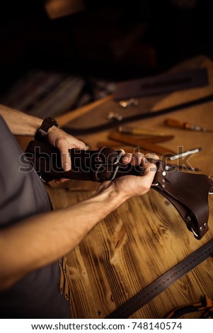Working process of the leather belt in the leather workshop. Man holding photographer's belt for camera. Tool on wooden background. Tanner in old tannery. Close up master's arm.
