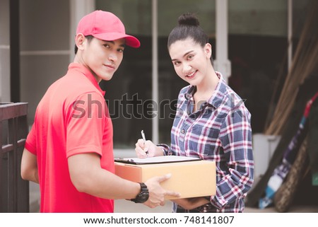 Young logistic career concept. Happy delivery man giving his package to customer at home. Taken in real house. Asian chinese fit man in red polo shirt and jeans with red hat in his early twenties.