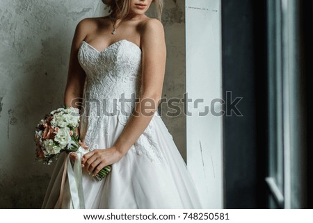 girl in a white wedding dress holding a bouquet of white flowers and greenery on the background of white brick wall