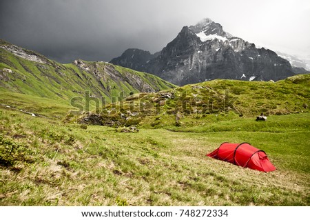 Red tent on a green and yellow grass, high alpine mountains in background. Dynamic and dramatic sky, storm clouds. Camping in a tent on an alpine meadow. Swiss alps.
