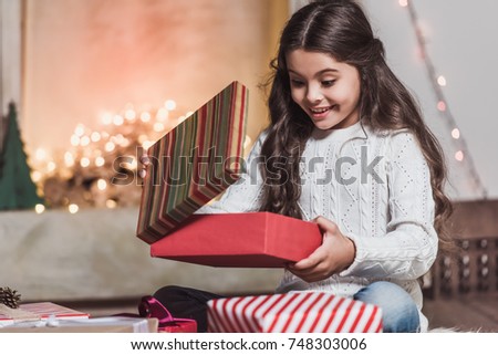 Merry Christmas and Happy New Year! Cute little girl is opening a present and smiling while sitting on the floor at home