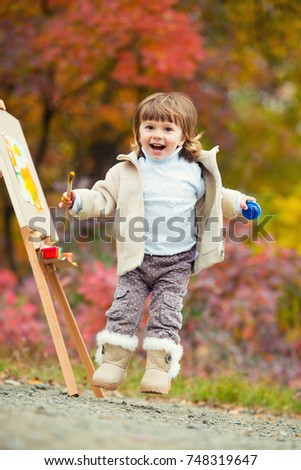 Cheerful little girl in a jump with a brush and paint, draws in the park of autumn leaves, painting of the Little Child, Child creativity.
