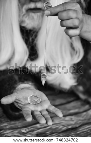 Old bearded man wizard with long white hair and beard holding blue gem stone and silver pendant for hypnosis on wooden background