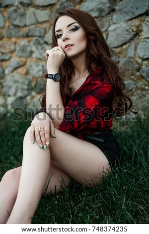 Beautiful young hipster girl posing and smiling near urban wall background in red plaid shirt, shorts, outdoors summer portrait.