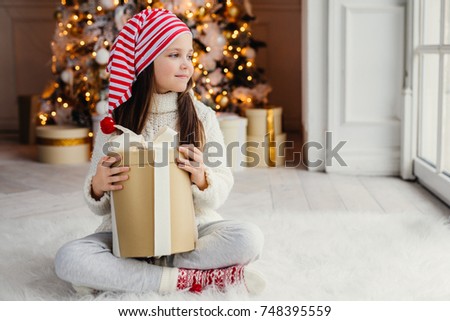 Adorable female child wears santa claus hat, warm clothes, hold wrapped gift box, sits against decorated New Year tree. Happy pretty kid looks aside out of window, sits crossed legs on floor