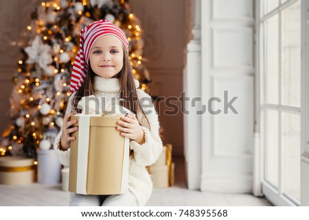 Indoor shot of pleasant looking small kid with blue charming eyes, wears santa hat, holds present in wrapped box, sits over decorated New Year background. Childhood, celebration concept