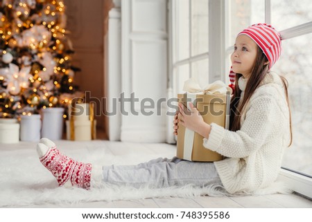 Sideways portrait of restful female child wears white sweater, trousers and warm socks, embraces wrapped gift, sits on floor in cozy room, admires New Year tree. Children and holidays concept