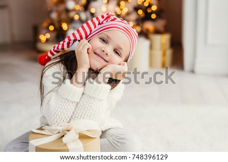 Beautiful small child poses in living room, leans at present gift, has happy expression, glad to receive surprise from parents, spends holidays in family circle. Merry Christmas and happy New Year
