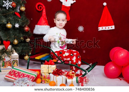 Funny kid sits on a sleigh next to a Christmas tree looking behind soap bubbles
