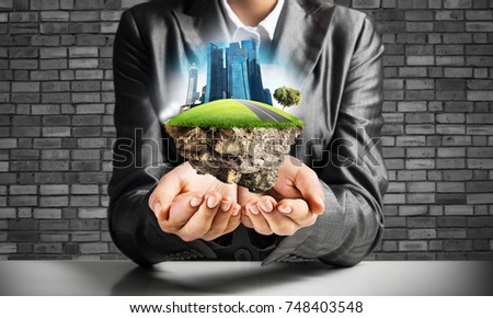 Businessman in suit keeping green island with skycraper city in hands with gray brick wall on background.