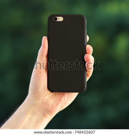 Smart phone on the blurred background of the park in a black plastic case back view. Smart phone in man's hand. Template of phone case Royalty-Free Stock Photo #748422607
