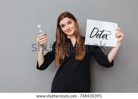 Portrait of pretty lady standing and holding postcard and bottle of water while dreamily looking in camera on gray background