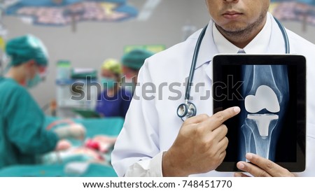 Male Doctor showing picture of Total knee replacement on tablet screen against blurred operating room. Medical technology concept.