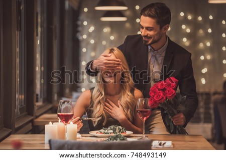 Handsome elegant man is holding roses and covering his girlfriend's eyes while making a surprise in restaurant, both are smiling Royalty-Free Stock Photo #748493149