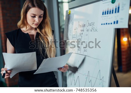 Female office worker working on her presentation standing near white board reading the report printed on paper Royalty-Free Stock Photo #748535356