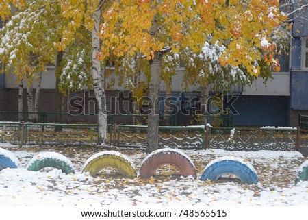 Decorative wheels from cars are buried in the ground on the playground in the city yard, colorful, covered with white snow. On the snowdrift yellow fallen leaves of trees. Autumn and winter background