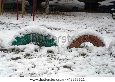 Decorative wheels from cars are buried in the ground on the playground in the city yard, colorful, covered with white snow. On the snowdrift yellow fallen leaves of trees. Autumn and winter background