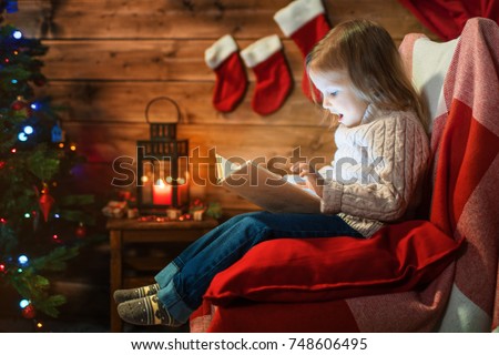 little girl at home with a christmas tree, gifts reading a book sitting in a red armchair