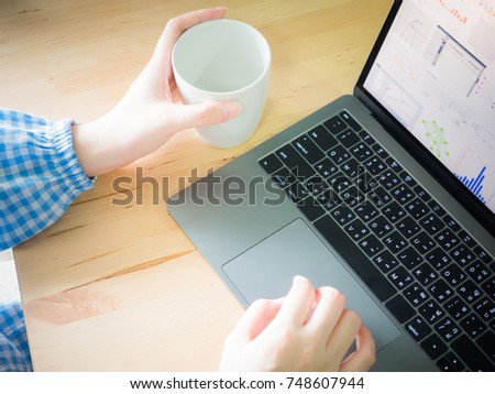 white hand with blue cloth of asian business woman hold cup and search data and check finance graph from internet for her business with laptop on wooden table background
